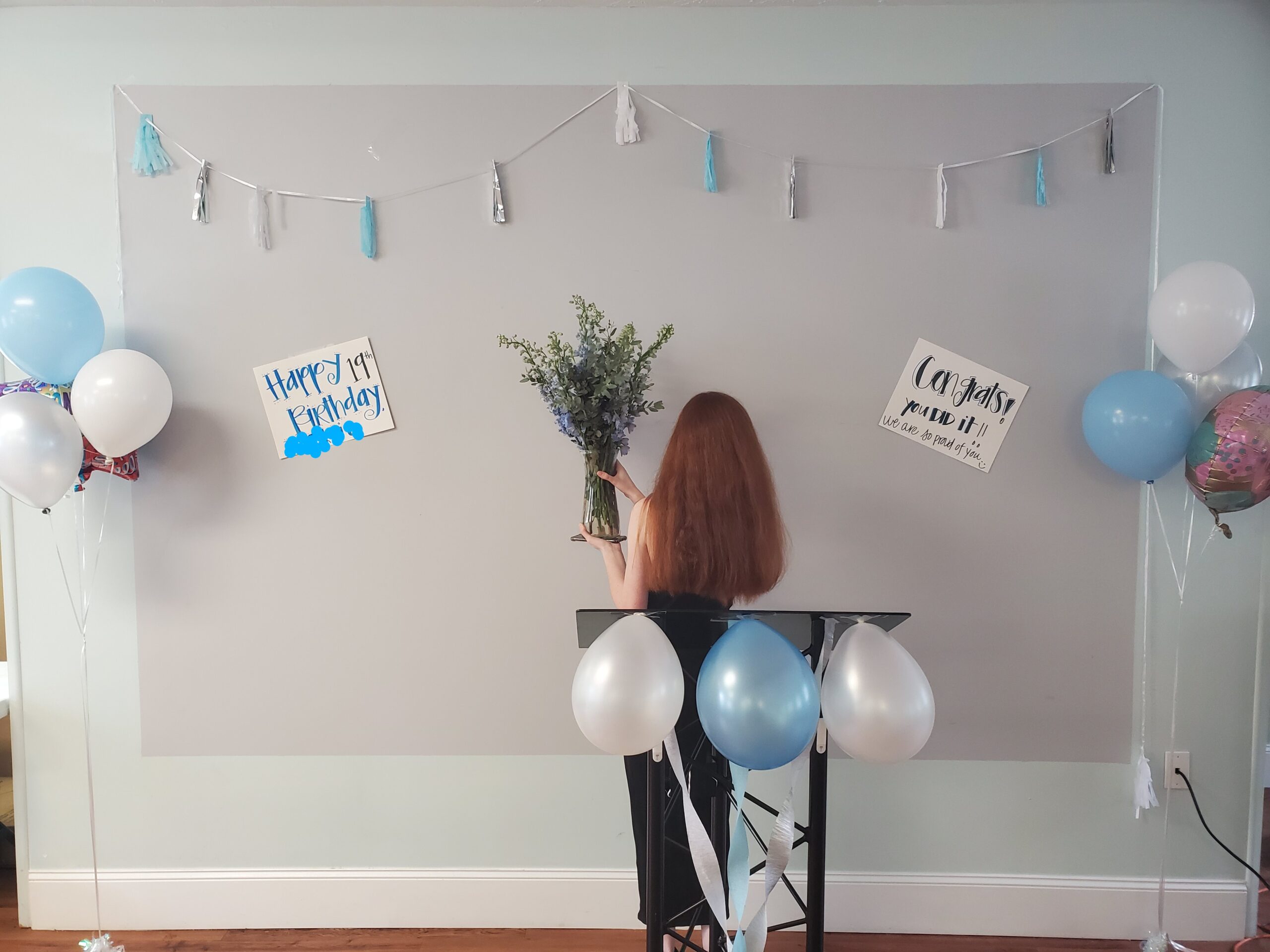Person with long red hair facing away, holding a vase of flowers at a blue-and-white birthday party with balloons and tassel garlands.