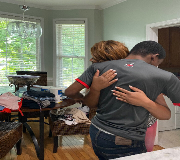 A man and woman stand in a room, facing away from the camera, with their arms around each other's backs. The woman's hands
